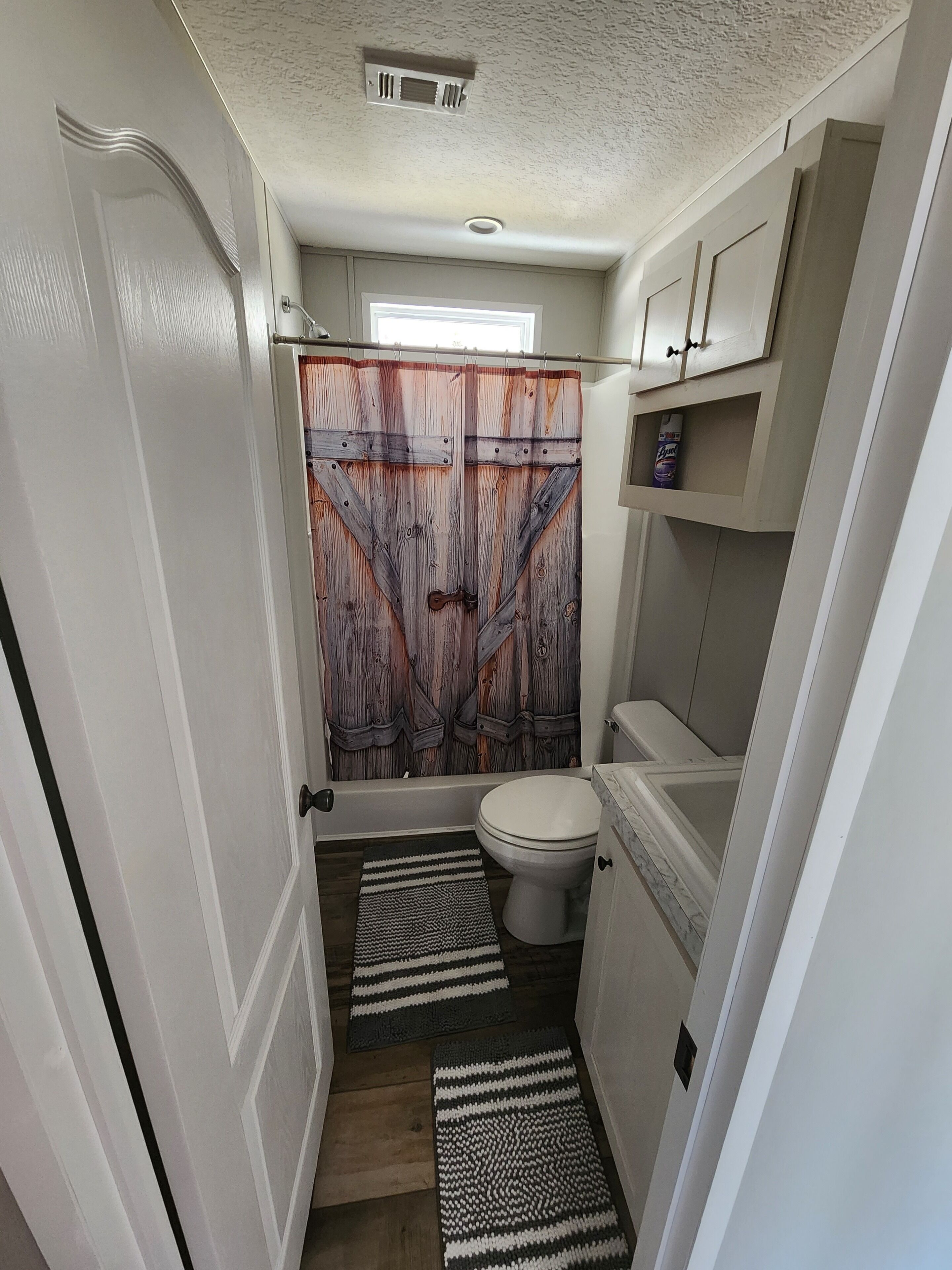 Une petite salle de bain avec un rideau de douche au style rustique, des toilettes, un lavabo et une petite étagère sur laquelle se trouve une bouteille.