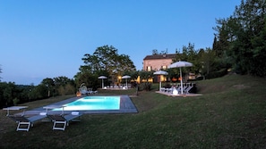 A swimming pool with lounge chairs and umbrellas, surrounded by a grassy area and trees.