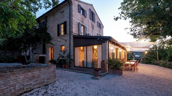 A two-story brick house with a patio area, outdoor seating, and a gravel driveway.