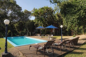 Zona de piscina con sillas de descanso y sombrillas, rodeada de árboles.