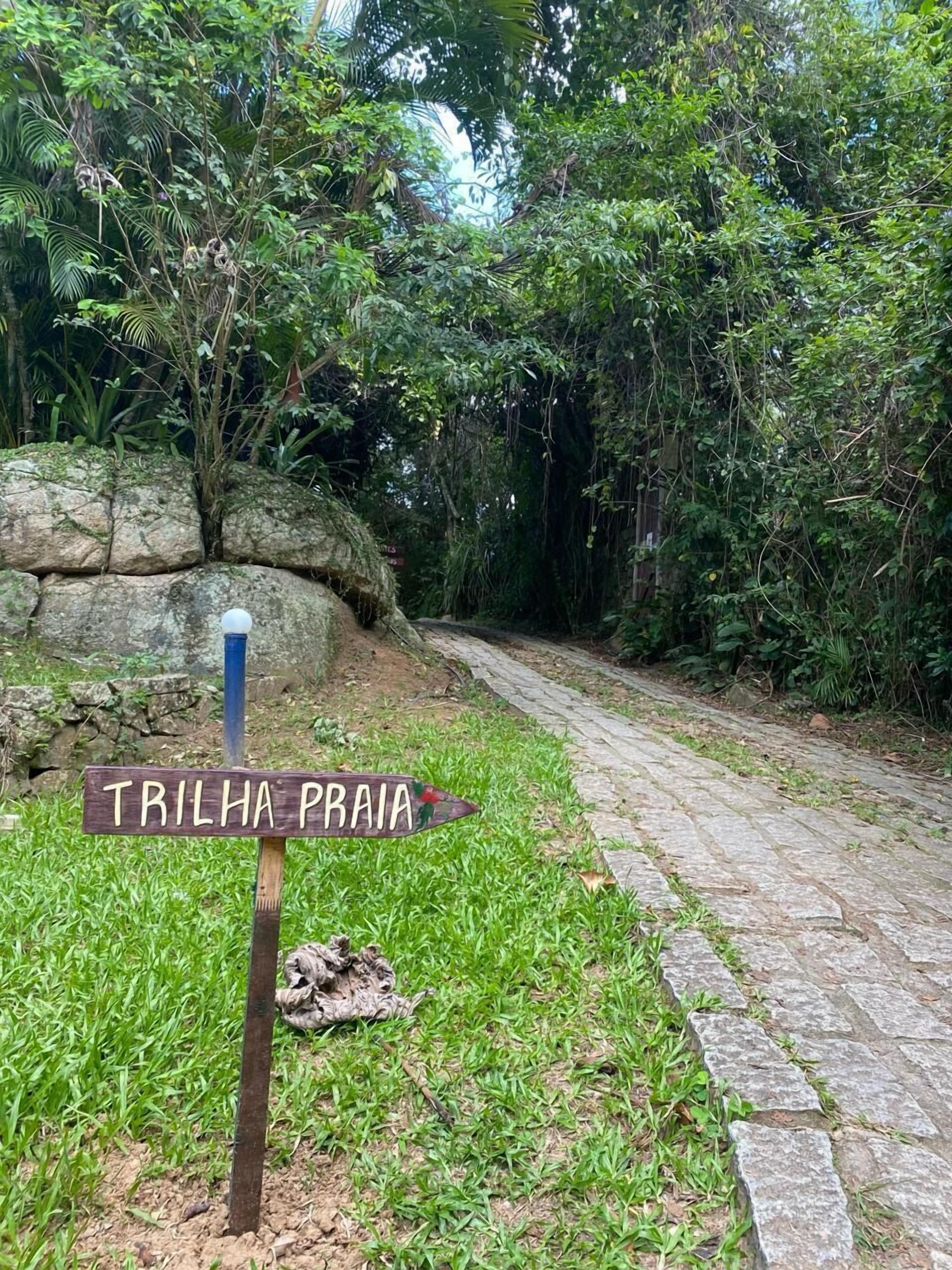 A stone pathway leading through a lush green area with a sign reading "TRILHA PRAIA".