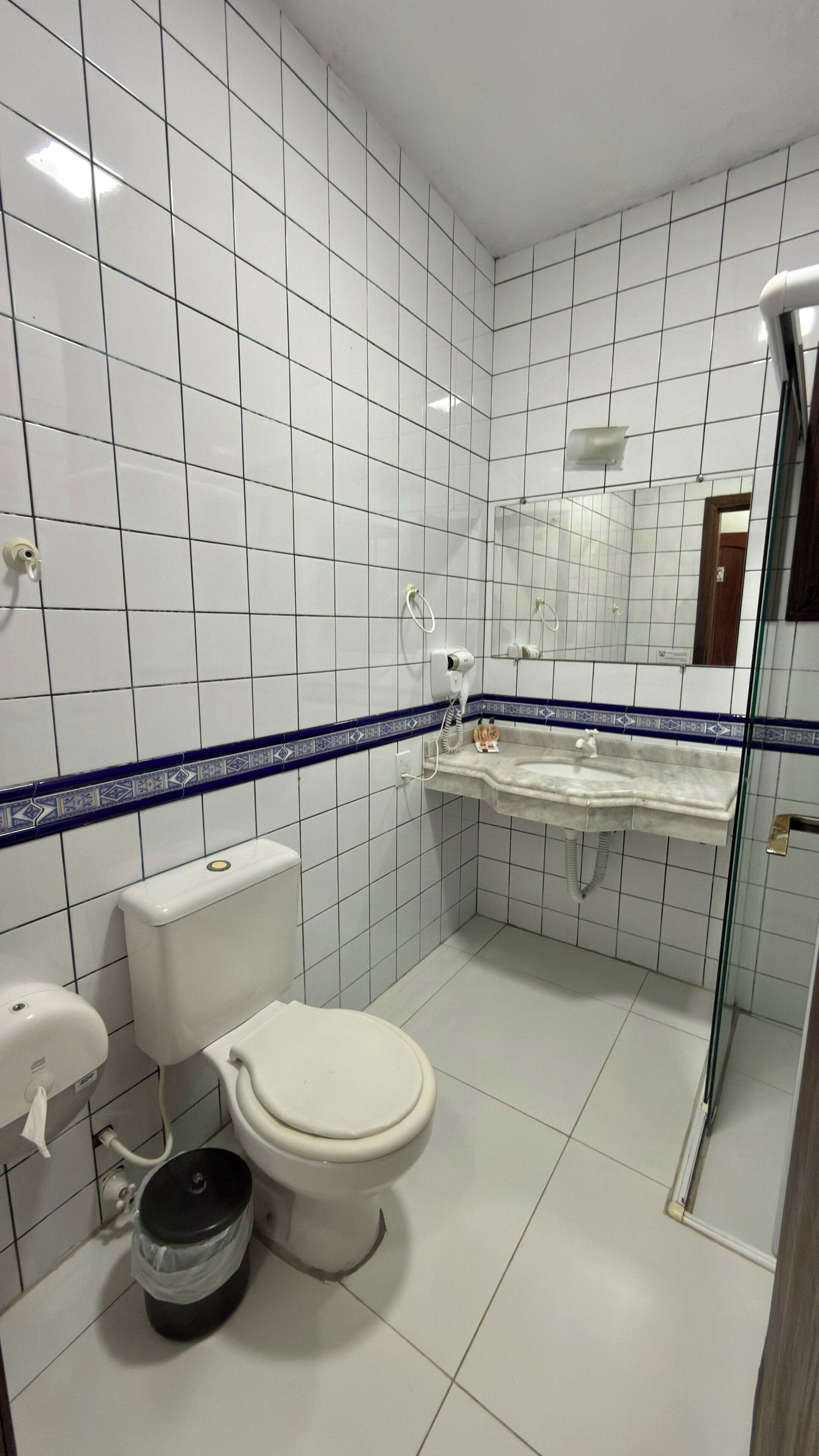 A bathroom with white tiles, a toilet, a sink, a shower, and a mirror.
