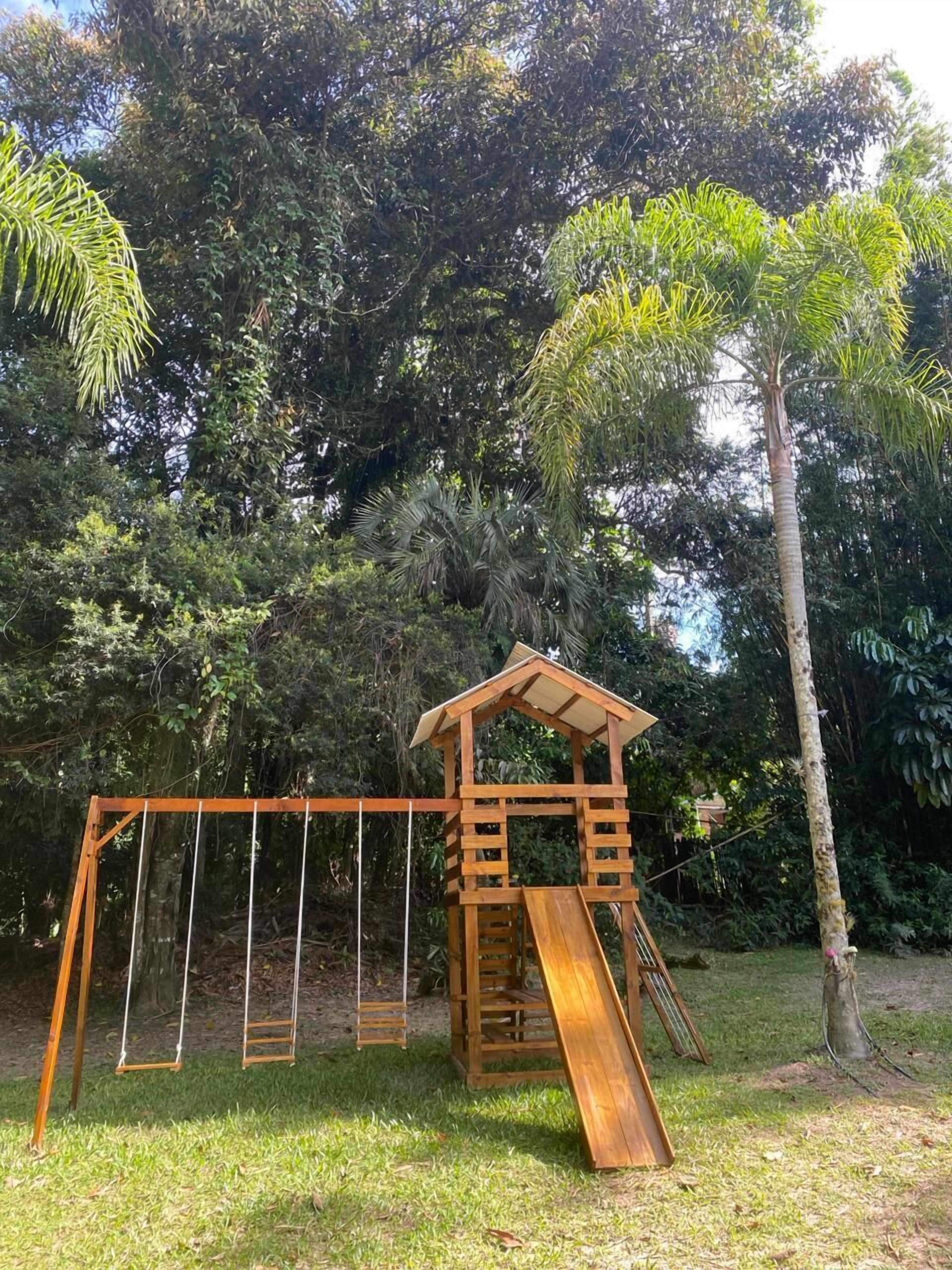 Um conjunto de baloiço de madeira com escorrega, rodeado por vegetação densa e árvores.