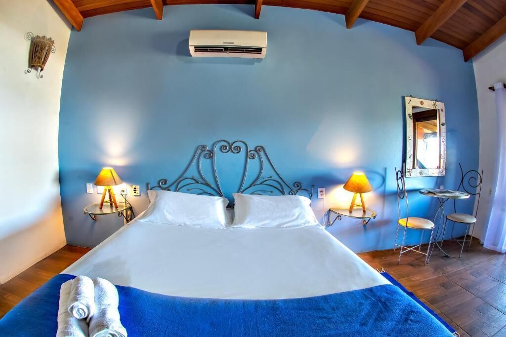 A bedroom with a large bed, two bedside lamps, a small table with chairs, a mirror, and an air conditioning unit.