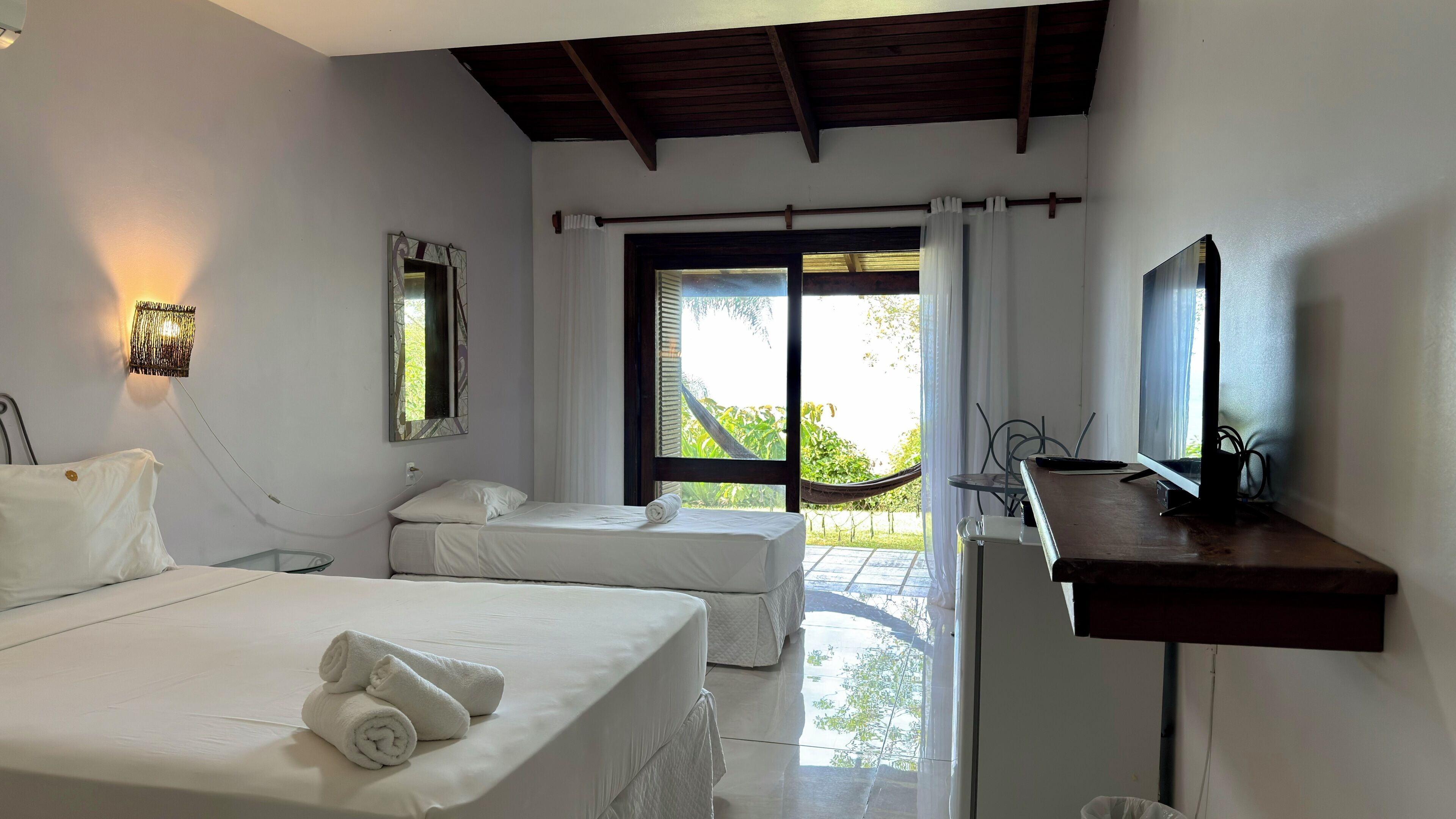 A hotel room with two beds, a desk, a TV, and a window with a view of greenery.