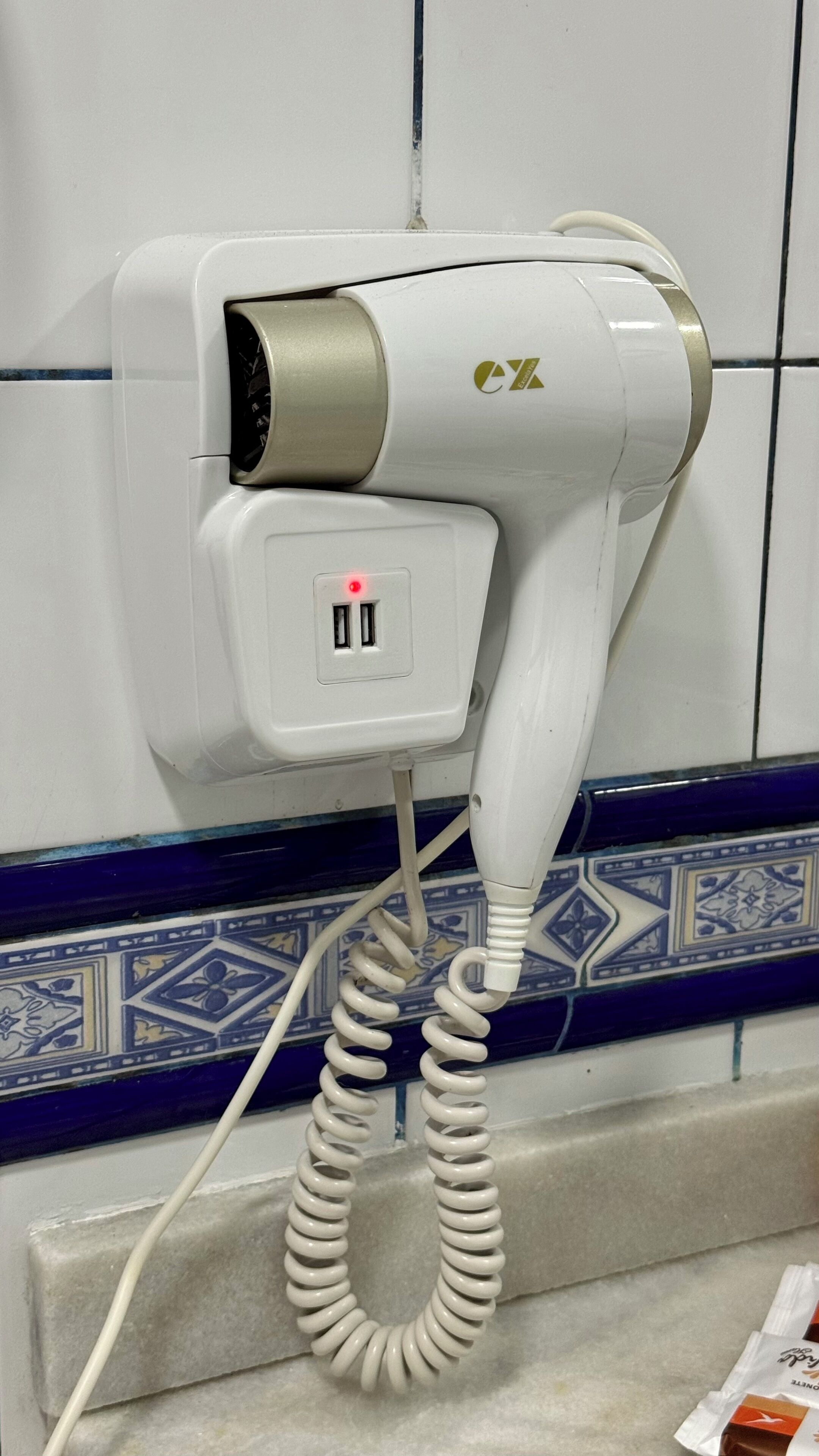 Un secador de pelo montado en la pared con un cable enrollado, un enchufe y un borde decorativo de azulejos.
