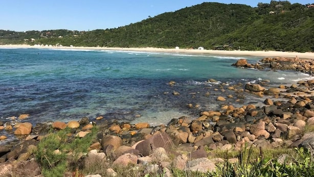 A rocky beach with clear water and a forested hill in the background. - Pousada Villa Agrifóglio (Imbituba)
