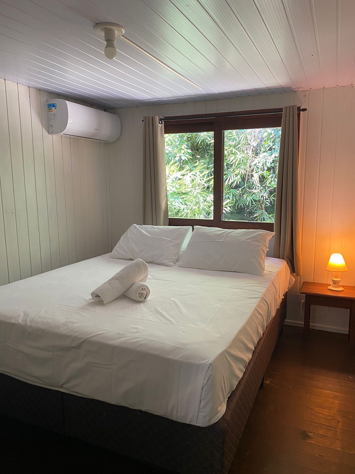 A neatly made bed with white linens and a rolled towel, a bedside lamp, and a window with curtains.