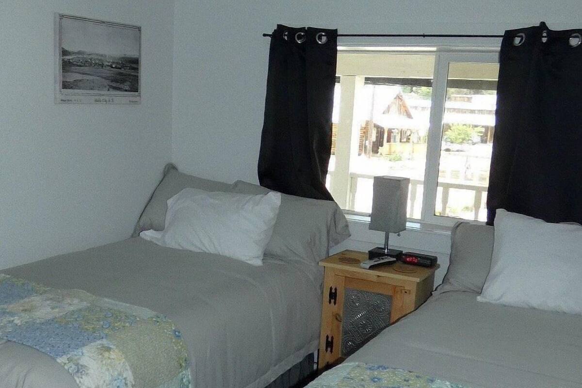 A room with two beds, a wooden bench, a nightstand with a lamp, and a window with curtains.