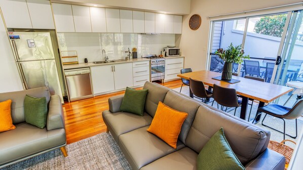 A modern kitchen with white cabinets, a stainless steel refrigerator, and a microwave. There is a dining area with a wooden table and chairs, and a sofa with orange and green cushions. - Kiali @ Cape Villas Dunsborough (Dunsborough)
