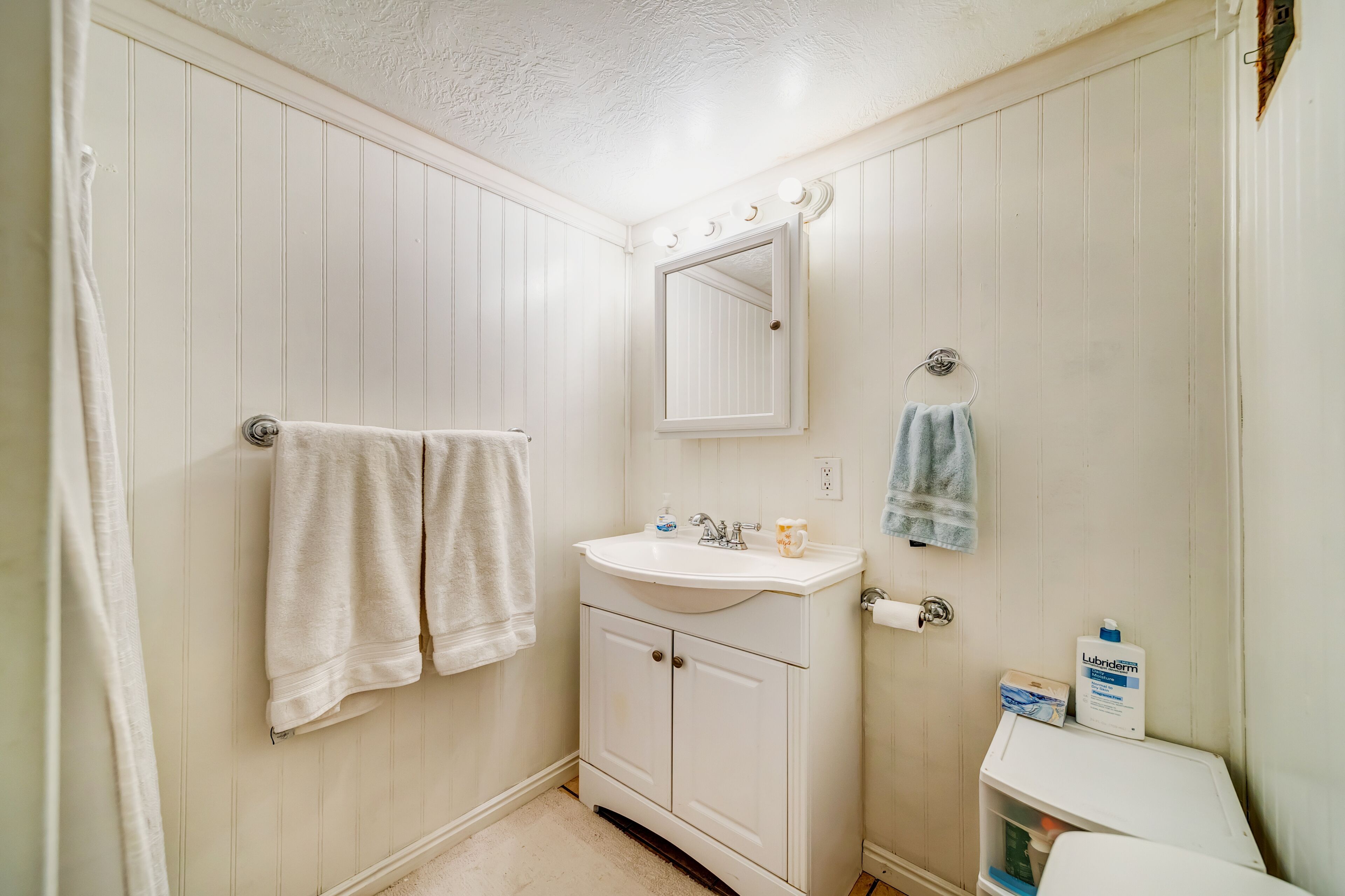 A bathroom with a white vanity, a mirror, a towel rack, and a bottle of Lubriderm.