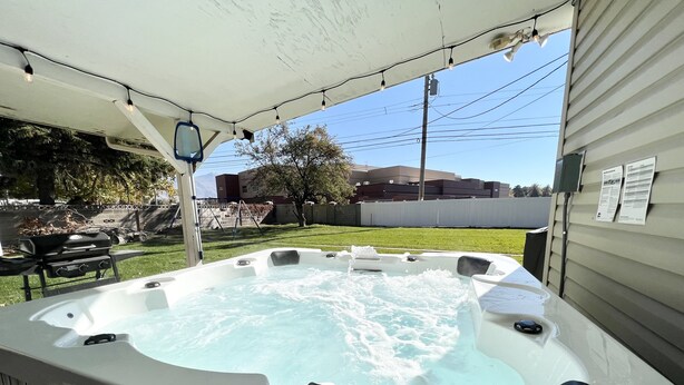 Un jacuzzi bajo una estructura cubierta, con una parrilla y mobiliario exterior al fondo.