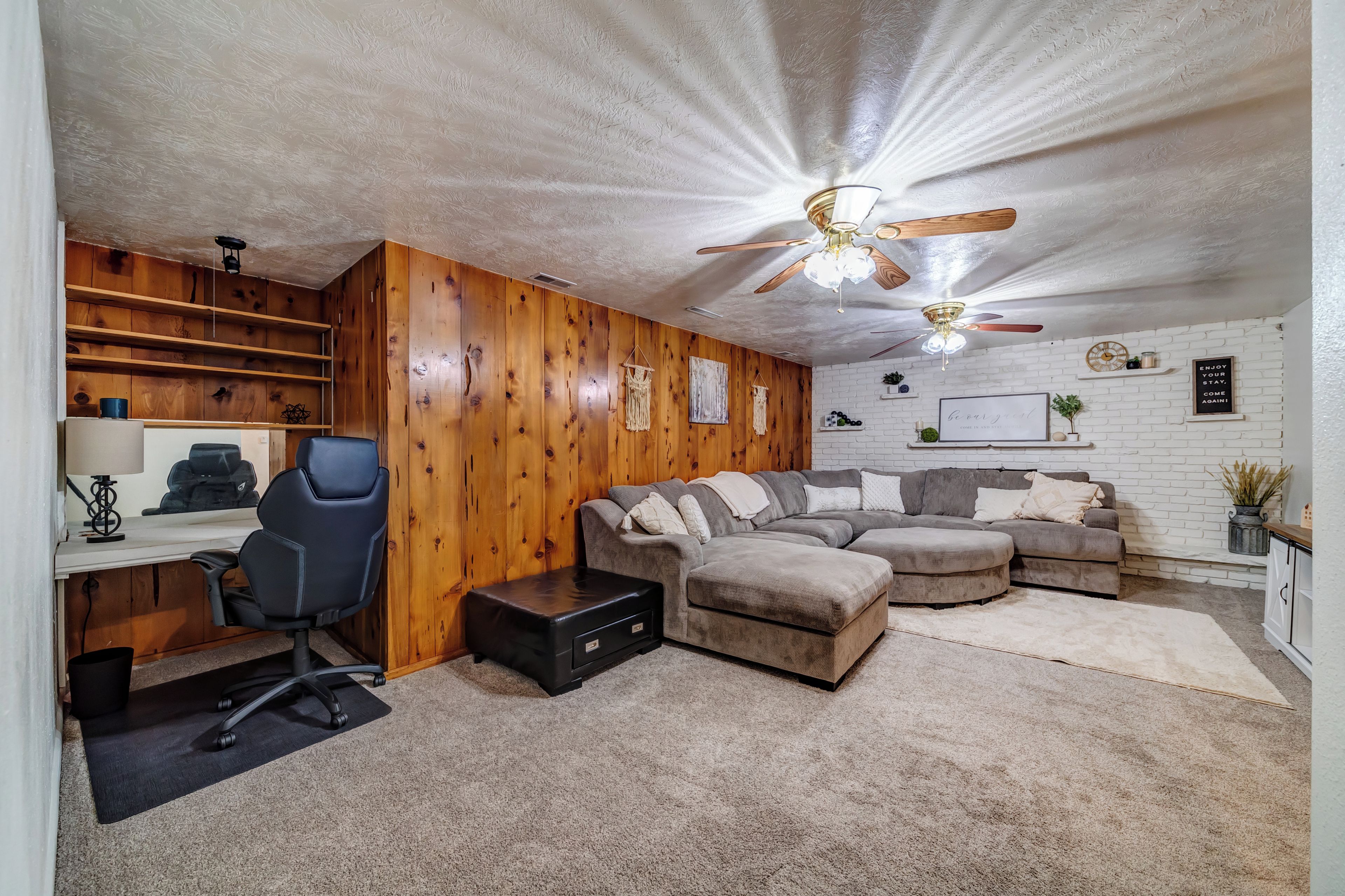 A room with a wooden wall, a gray sectional sofa, a black office chair, a desk with a computer, and a ceiling fan.