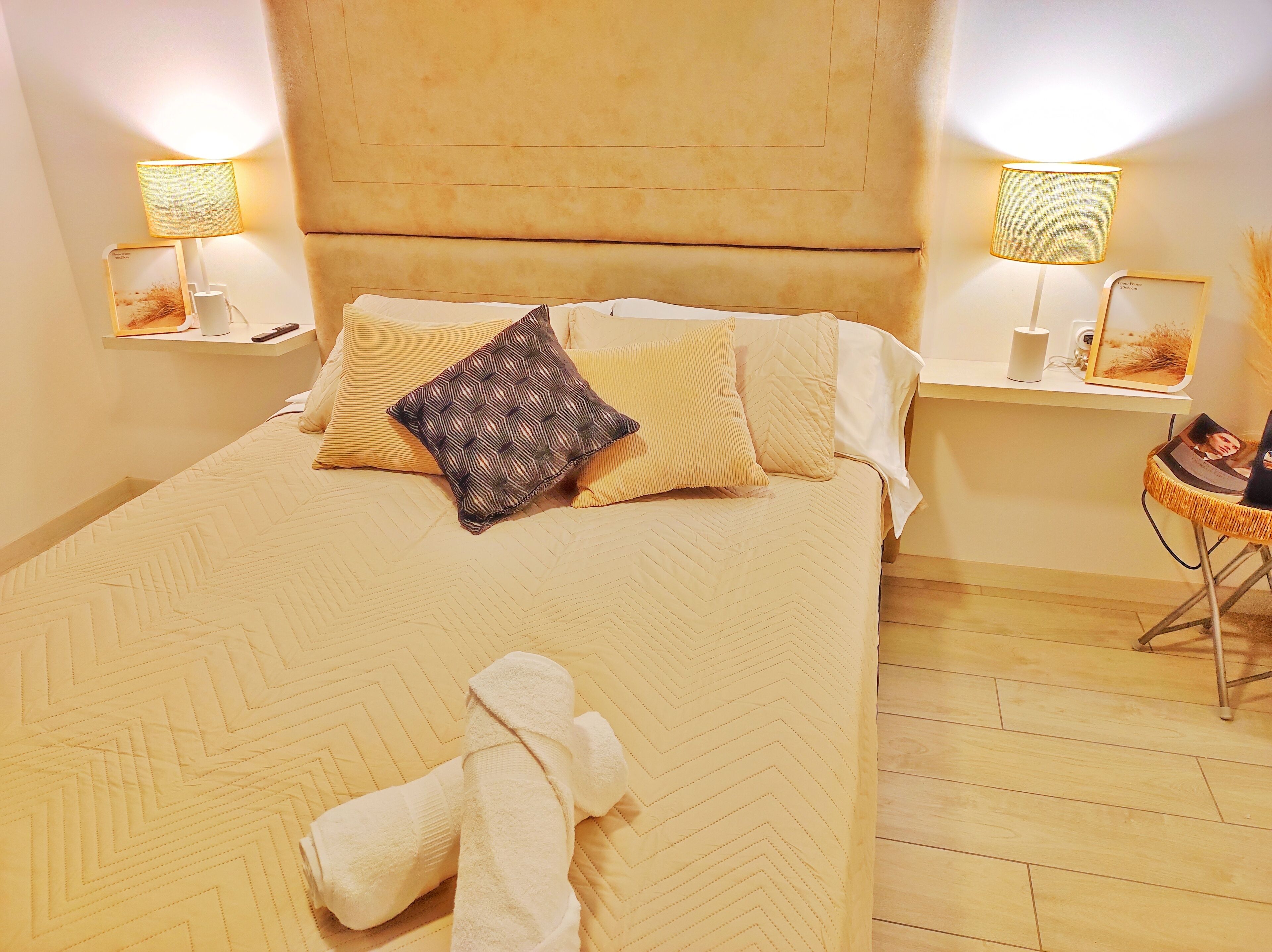 A neatly made bed with a patterned bedspread, two pillows, and a folded towel on the floor. There are two bedside tables with lamps and a small wicker chair.