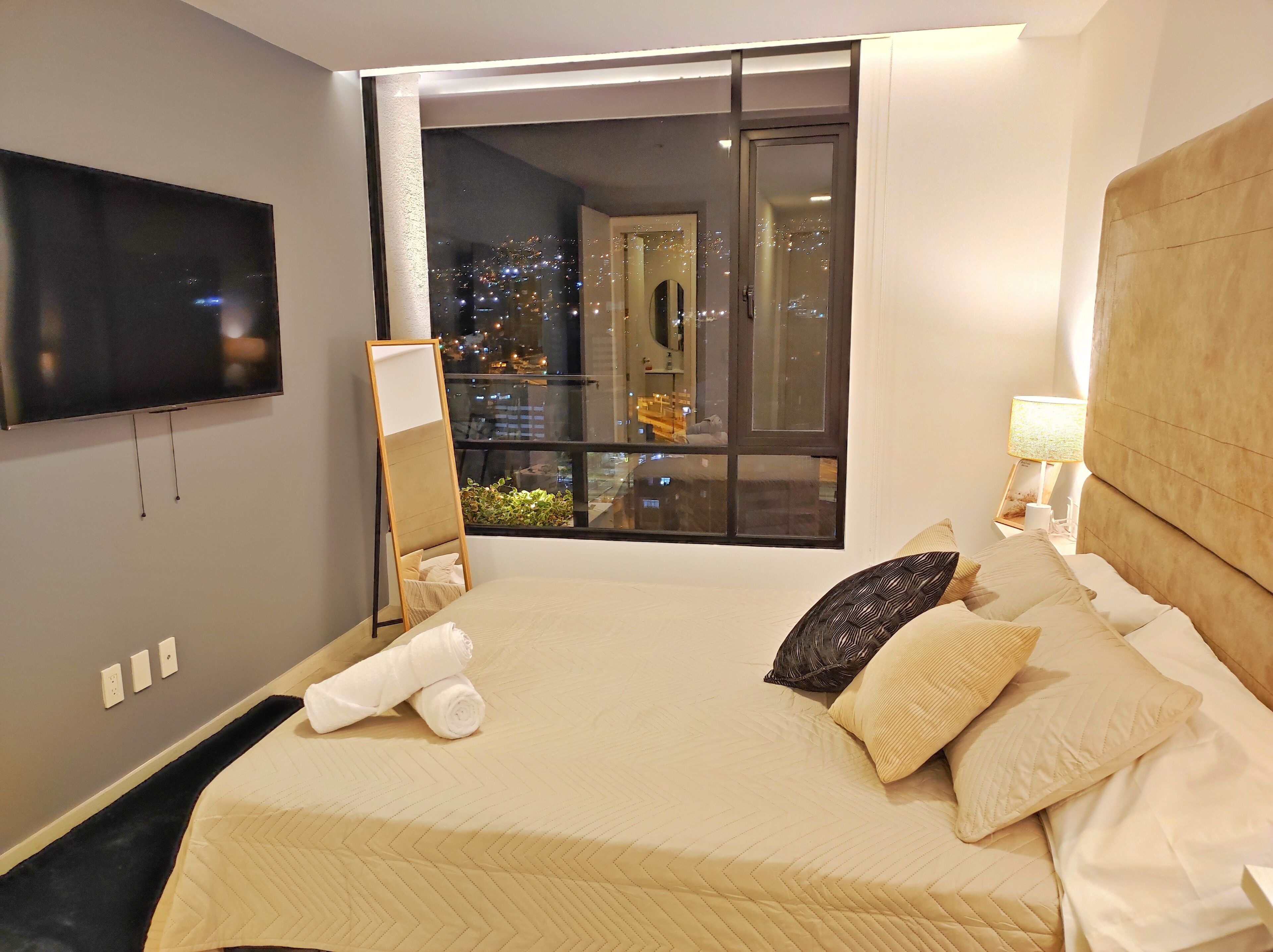 Une chambre d’hôtel dotée d’un grand lit, d’un téléviseur à écran plat fixé au mur, d’une fenêtre offrant une vue sur la ville la nuit et d’une table de chevet avec une lampe.