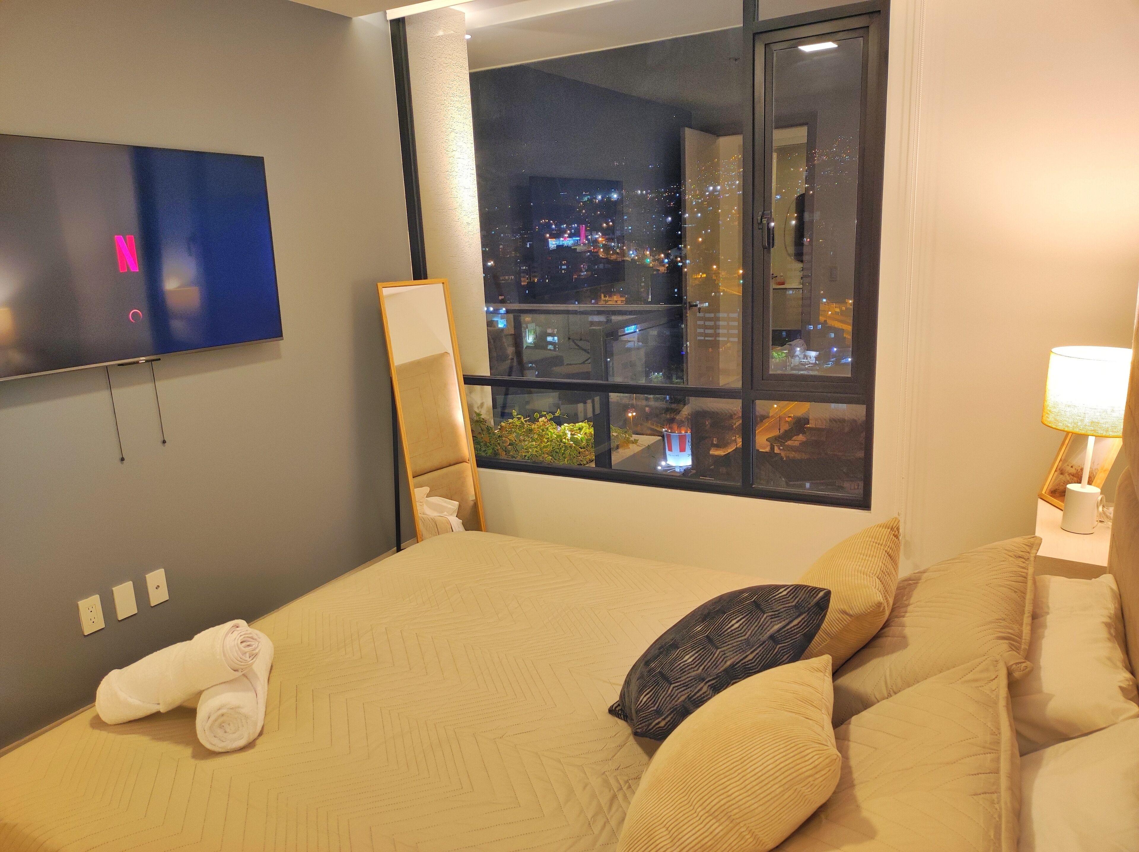 A hotel room with a bed, a TV mounted on the wall, a window with a city view, and a nightstand with a lamp.