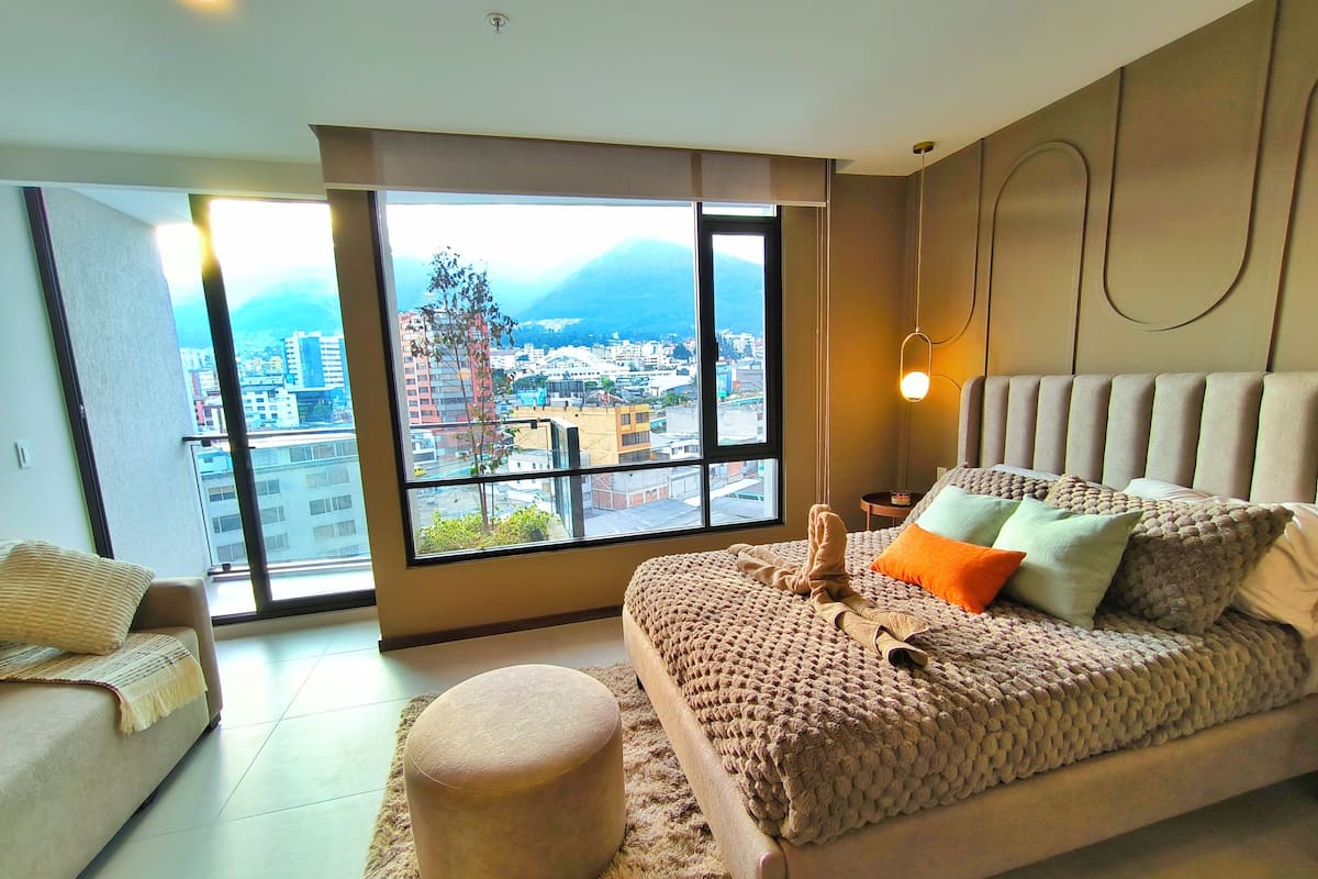 A modern bedroom with a large window, a bed with multiple pillows, a round ottoman, and a view of a cityscape.