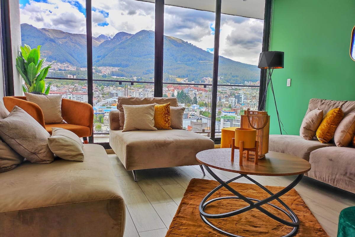 A modern living room with a large window offering a cityscape and mountains view.