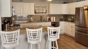 A modern kitchen with white cabinets, a central island with bar stools, a stainless steel refrigerator, and a decorative wall hanging. - WW Condo 1.5 blks to beach and boardwalk. Walk to shops, restaurants and CC. (Wildwood)