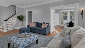A spacious living room with a grey sofa, a blue sofa, and a patterned ottoman. There is a staircase with white railings and a potted plant. - Spacious Home on Gervais | 4BR 2BA | Downtown (Columbia)