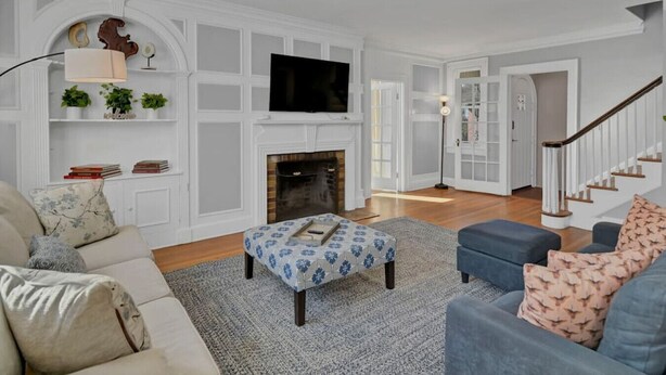 A living room with a fireplace, a flat-screen TV mounted above it, a sofa, an ottoman, and a staircase leading to another room. - Spacious Home on Gervais | 4BR 2BA | Downtown (Columbia)