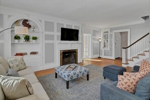A living room with a fireplace, a flat-screen TV mounted above it, a sofa, an ottoman, and a staircase leading to another room. - Spacious Home on Gervais | 4BR 2BA | Downtown (Columbia)
