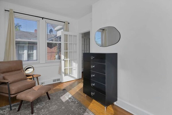 Een kamer met een bruine leren fauteuil, een zwarte dressoir met lades, een ronde spiegel en een raam met jaloezieën.