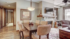 A dining area with a wooden table and patterned chairs, a fireplace with stone surround, and a television mounted above it. - Waterhouse 5109 (Breckenridge)