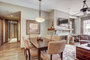 A dining area with a wooden table and patterned chairs, a fireplace with stone surround, and a television mounted above it. - Waterhouse 5109 (Breckenridge)