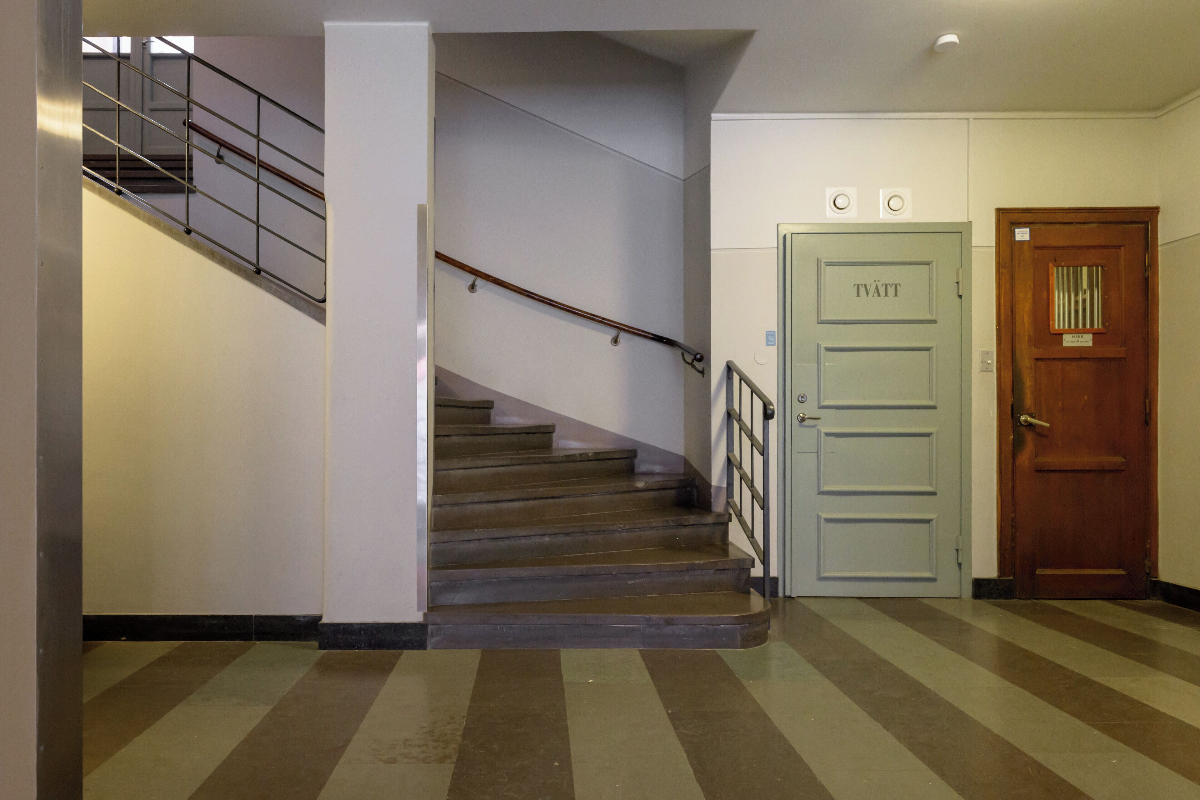 A staircase with metal railings, a wooden door labeled "TVATT," and a striped floor pattern.