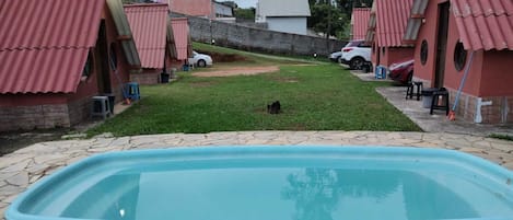 A swimming pool with a stone deck, surrounded by a grassy area and a few buildings with red roofs.