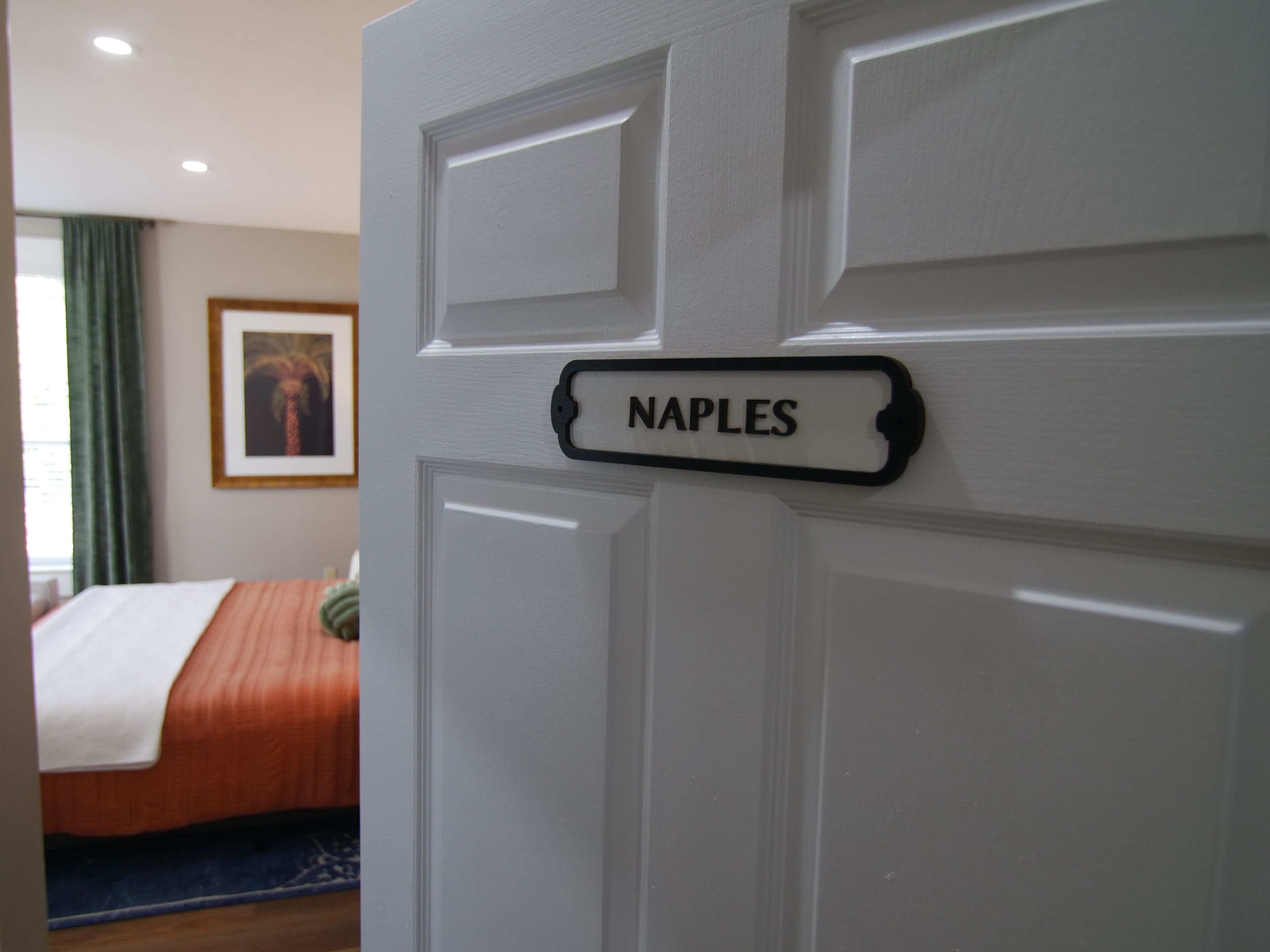 Hotelkamer met een bed, een ingelijst schilderij aan de muur en een deur met een naambordje waarop 