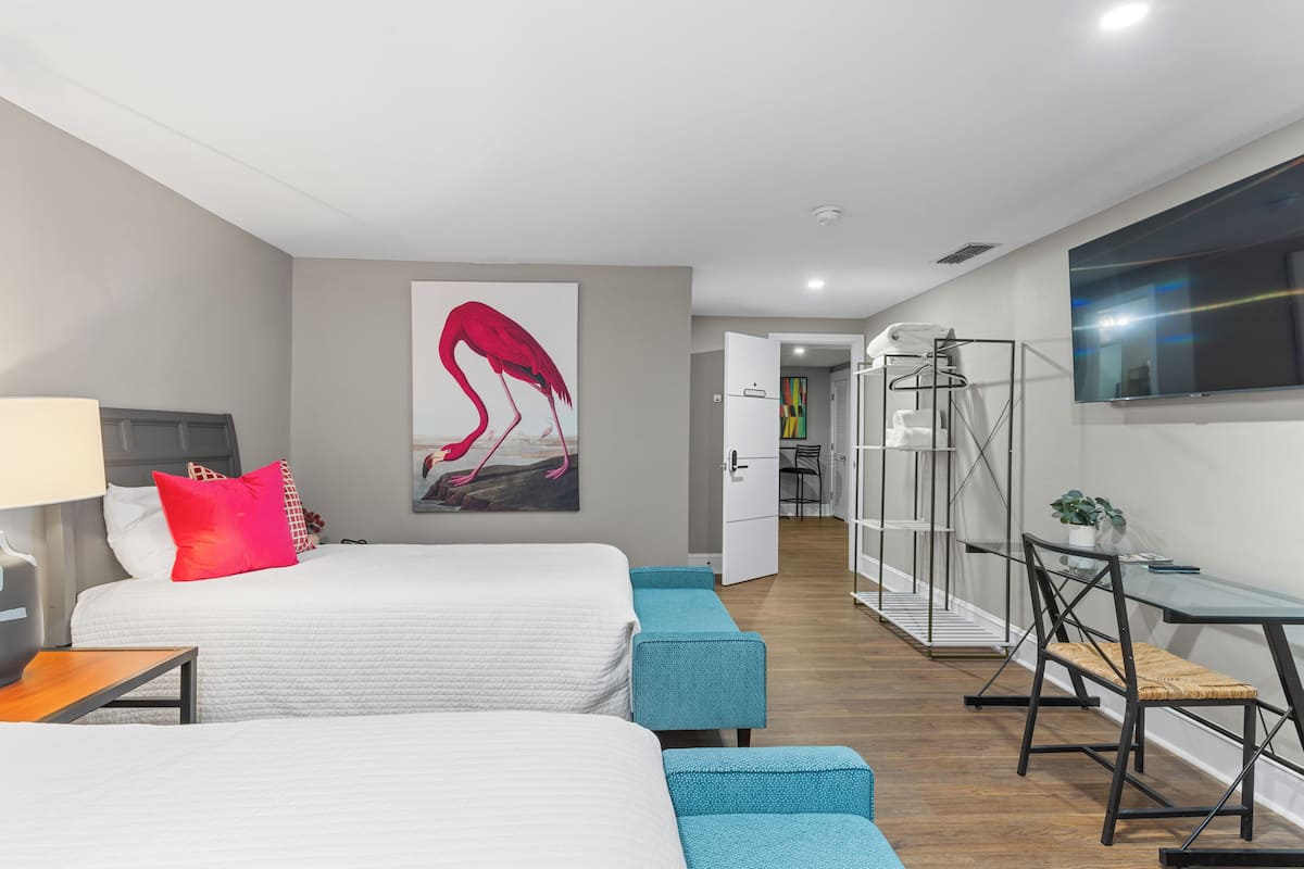 A modern hotel room with two beds, a desk, a chair, a TV, and a painting on the wall.