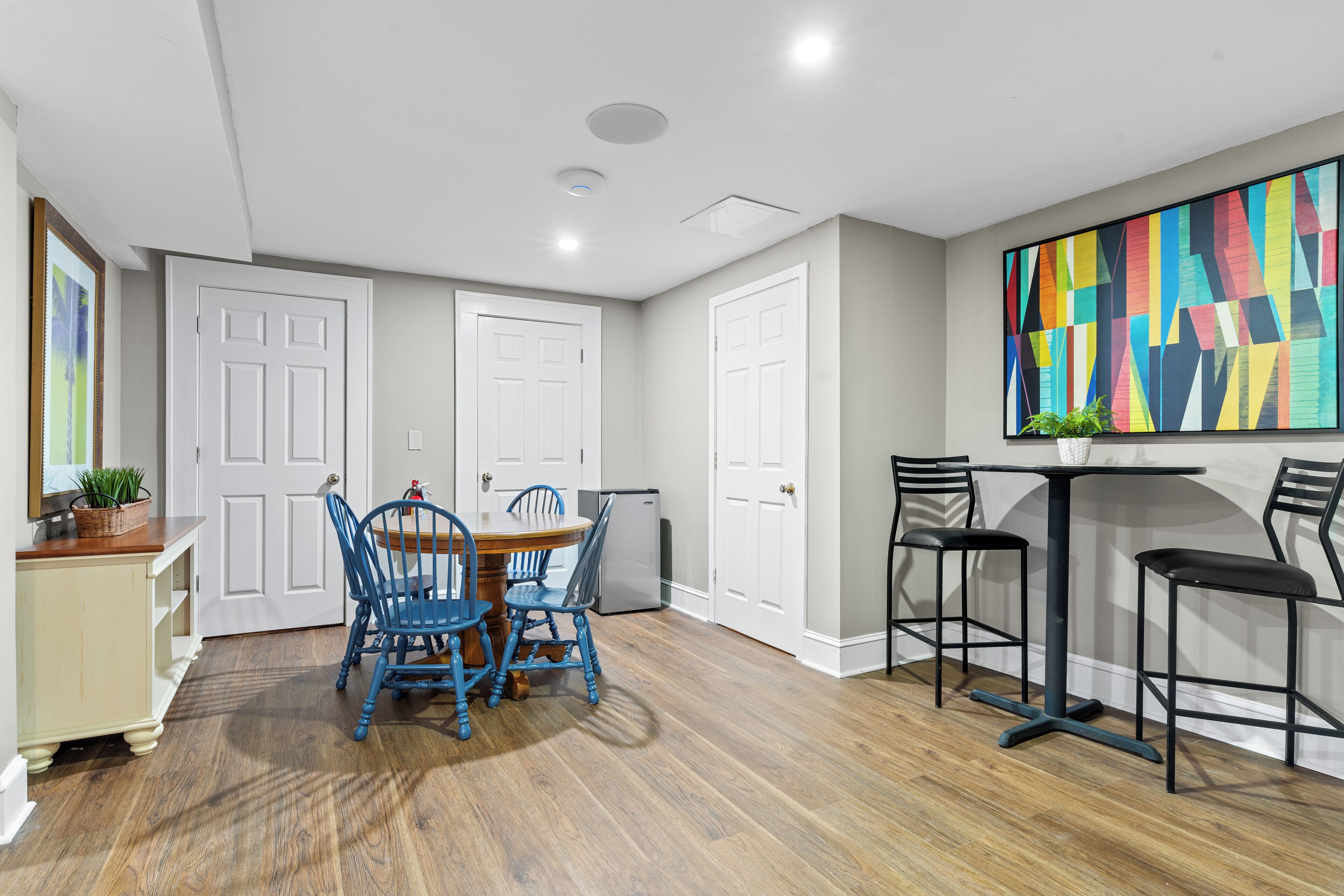 A dining area with a wooden table and blue chairs, a bar with stools, and a colorful abstract painting on the wall.