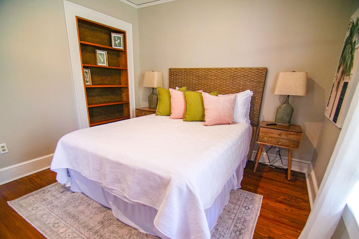 A bedroom with a large bed, a wooden headboard, a bedside table with a lamp, a nightstand, a built-in bookshelf, and a patterned rug.