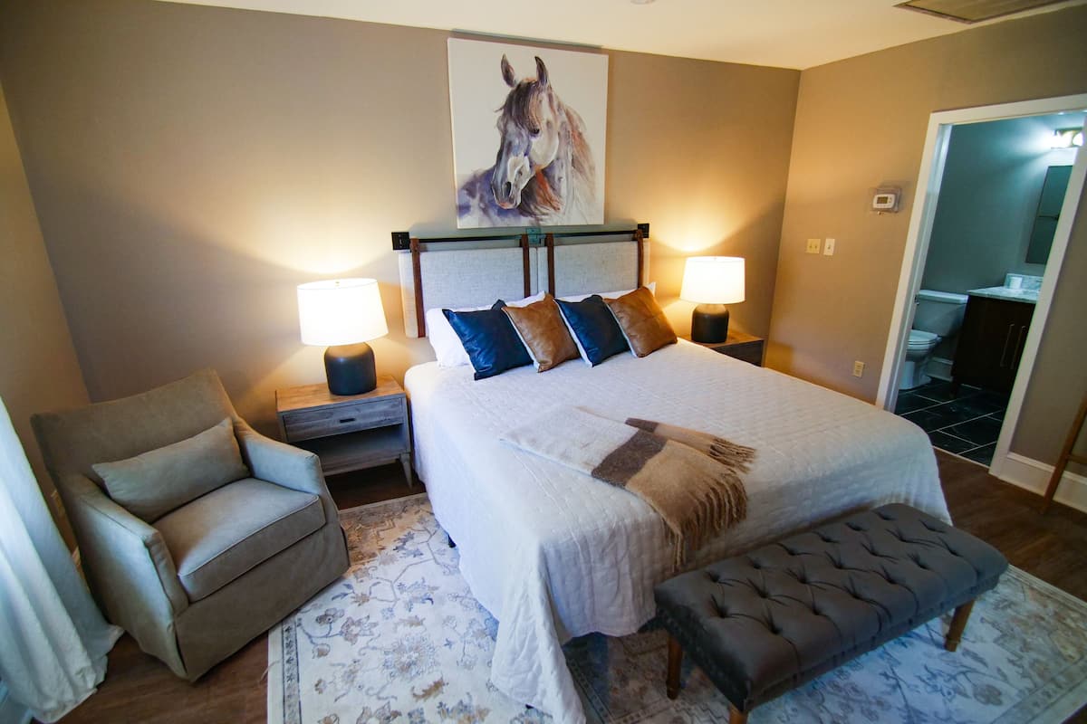 A hotel room with a large bed, two bedside tables with lamps, a chair, a bench, a rug, and a painting on the wall.
