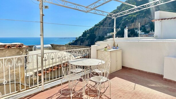 A terrace with a white metal railing, a round table with chairs, and a view of the sea and a mountain.