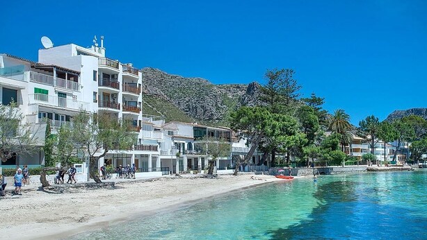 A coastal building with balconies overlooking a clear blue sea, surrounded by trees and a mountainous backdrop.