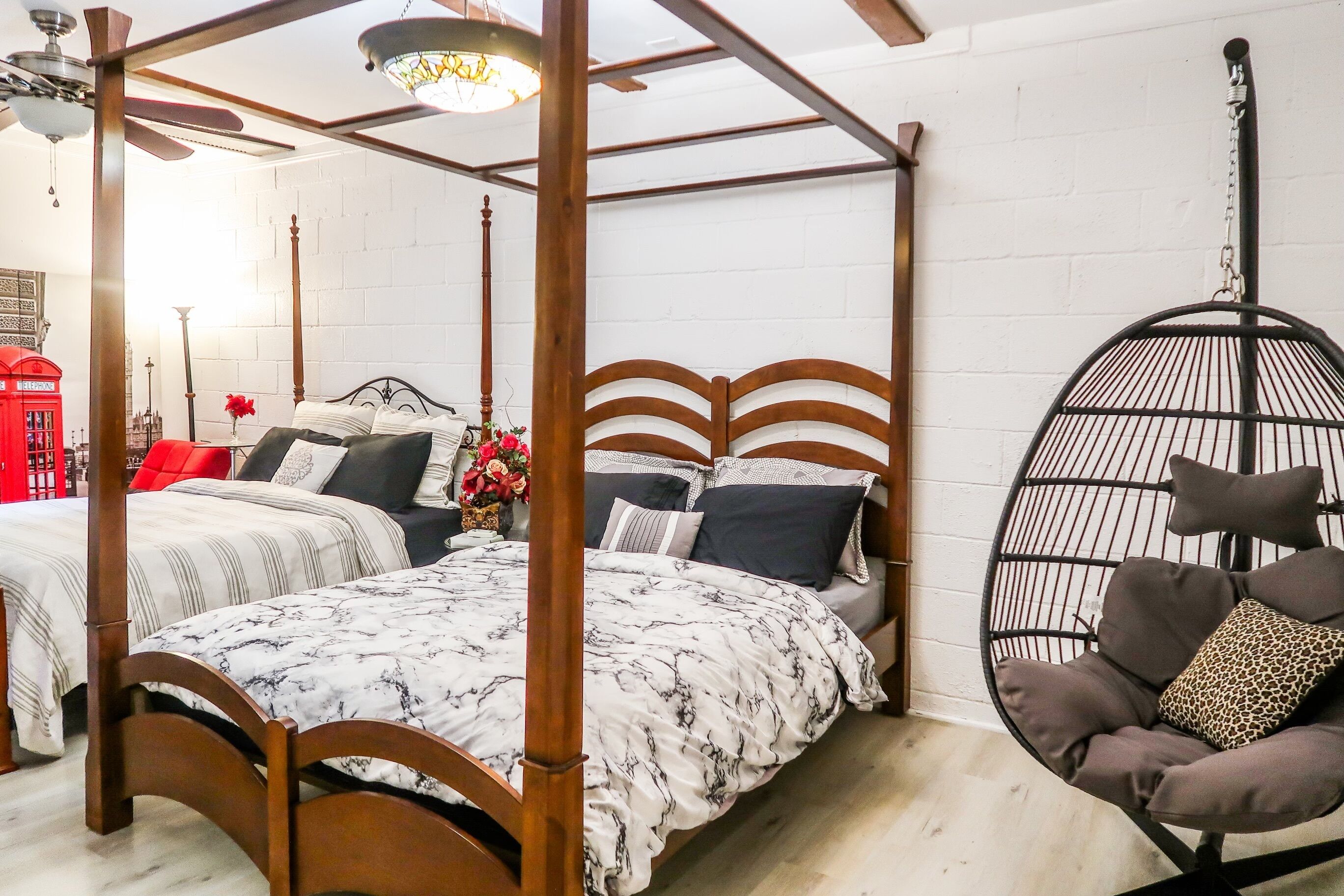 Un lit à baldaquin avec une couvre-lit à motifs noirs et blancs, un fauteuil suspendu avec des coussins, et une cabine téléphonique rouge en arrière-plan.