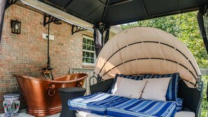 A covered outdoor seating area with a cushioned bench, a copper bucket, and a lantern against a brick wall.
