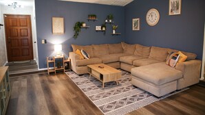 Un salon comprenant un canapé d’angle, une table basse, un tapis à motifs et des étagères murales ornées de plantes.