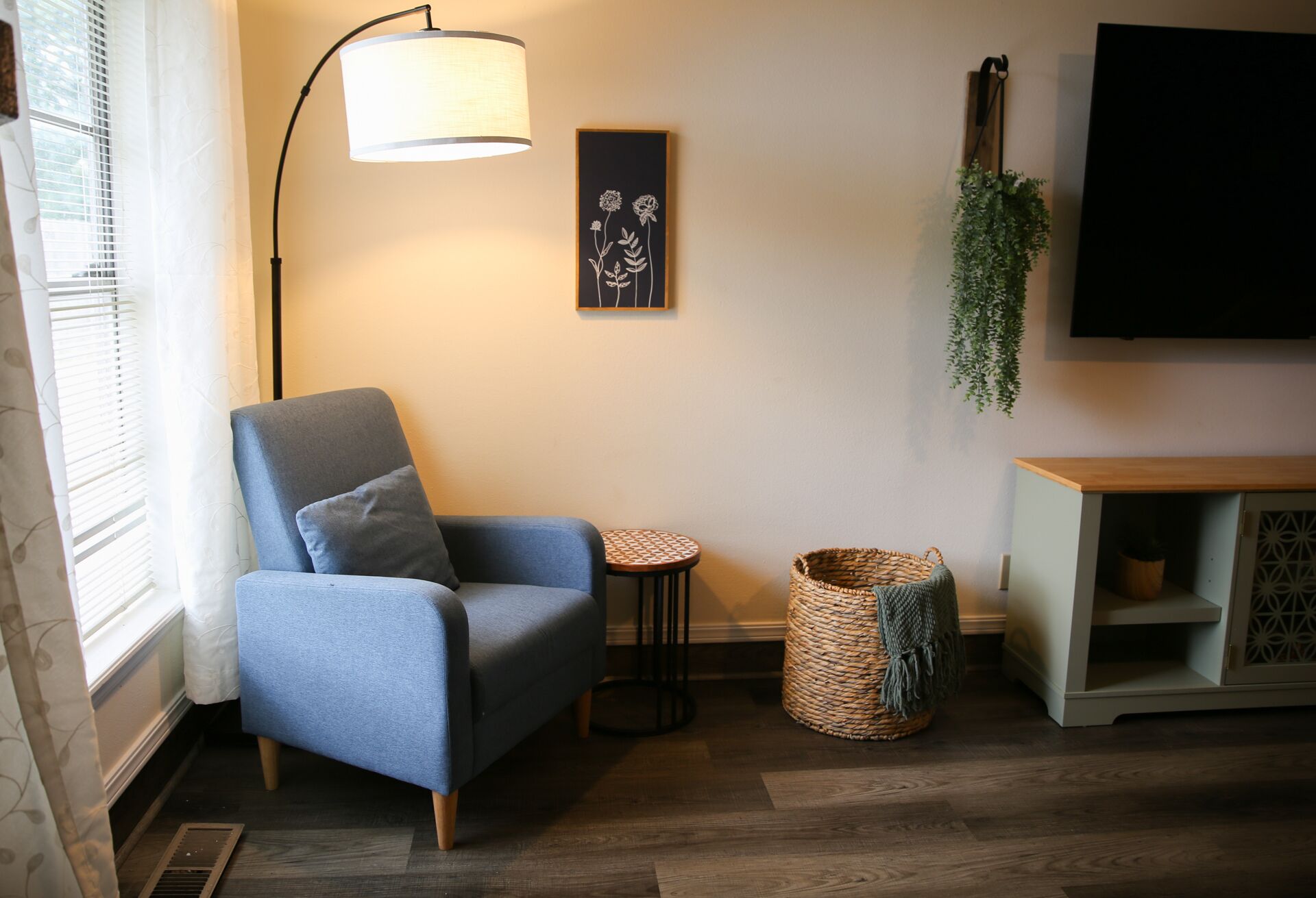 Ein Wohnzimmer mit blauem Sessel, einem kleinen runden Tisch, einem gewebten Korb, einer Wandpflanze und einem Fernseher.