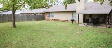 A backyard with a lawn, a wooden fence, a stone wall, and a covered patio area with chairs and a table.