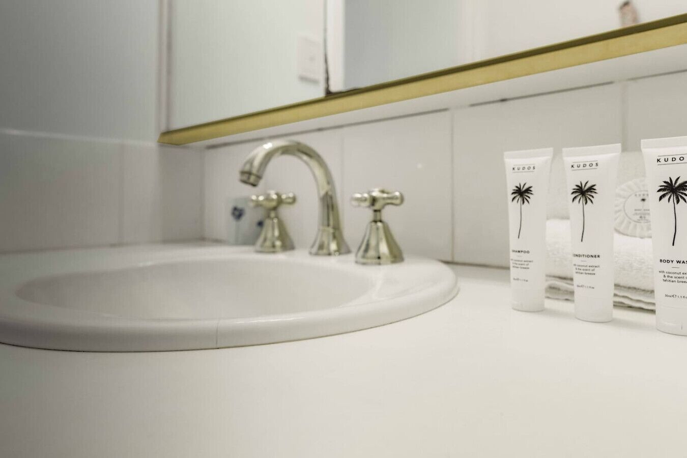 Ein Badezimmer mit einem weißen Waschbecken, zwei Wasserhähnen und drei Flaschen des Produkts KUDOS auf der Ablagefläche.