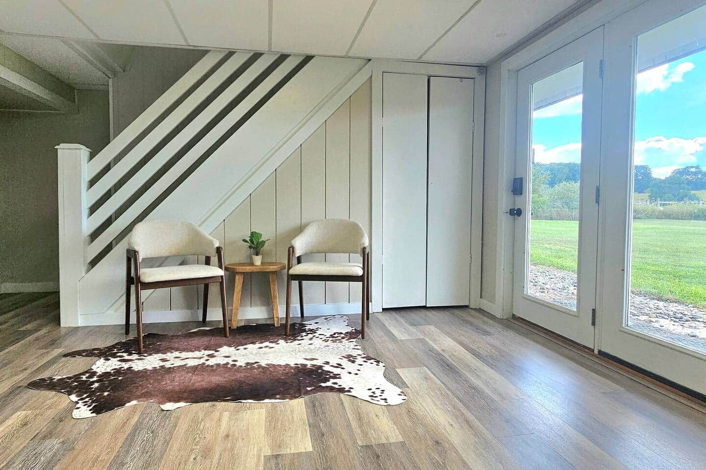 Ein Zimmer mit Holzboden, einem Kuhfellteppich, zwei Stühlen, einem kleinen Tisch mit Pflanze und einer Glastür zum Garten.