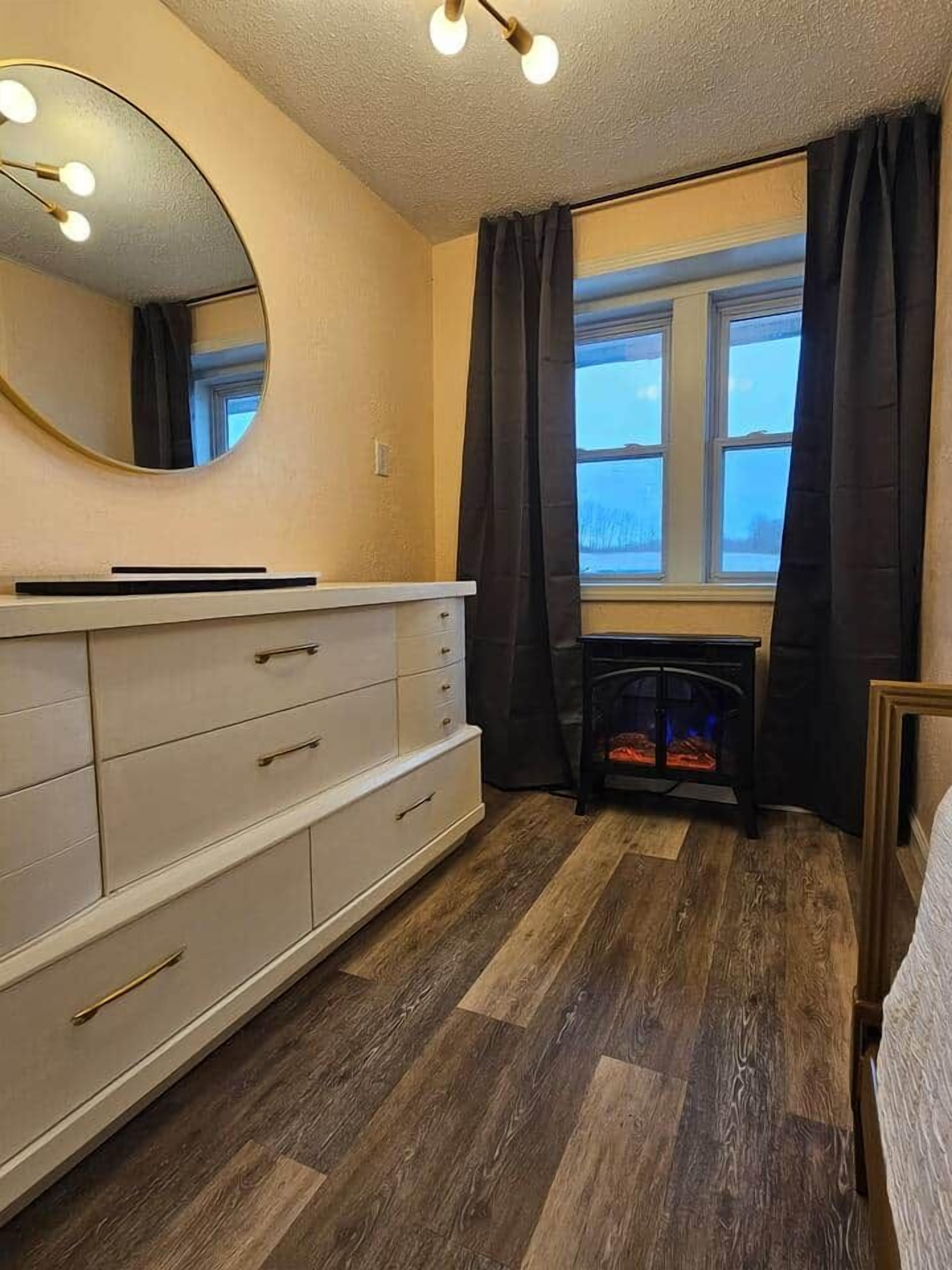 Un dormitorio con cómoda, espejo, ventana con cortinas y chimenea.