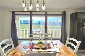 Un comedor con mesa de madera, sillas blancas, lámpara colgante y vista a un patio cercado a través de grandes ventanales.