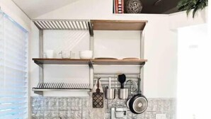 A compact kitchen with a built-in dishwasher, a coffee machine, and a kettle on the countertop. There are shelves above the sink for storage, and a window with blinds on the left. - Cozy Tiny studio 20Min to DwTn SF & 7min to SFO (South San Francisco)