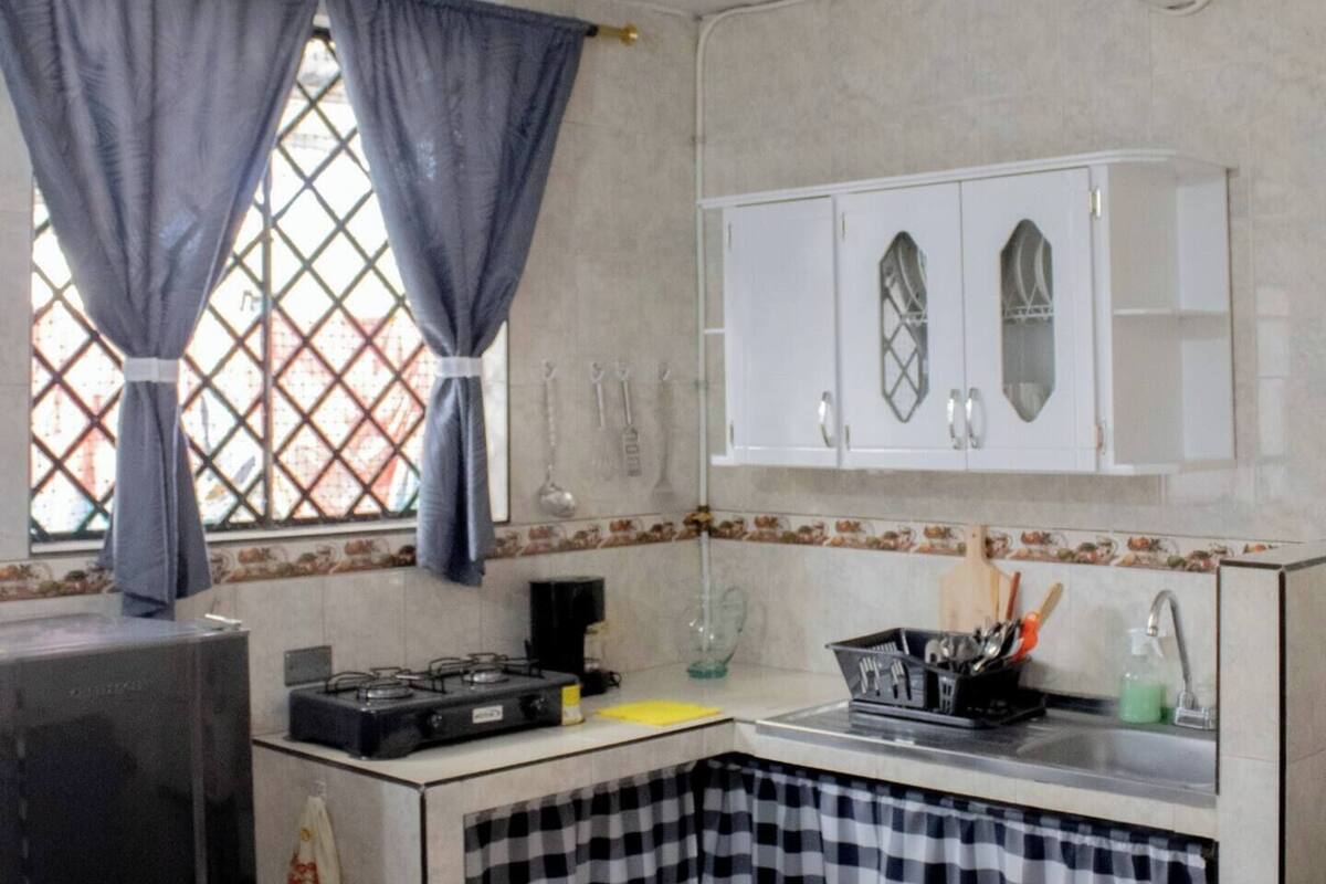 Une cuisine de taille réduite, équipée d’une cuisinière à gaz, d’un réfrigérateur et d’un évier. On y trouve des placards au-dessus du plan de travail et une fenêtre avec des rideaux.