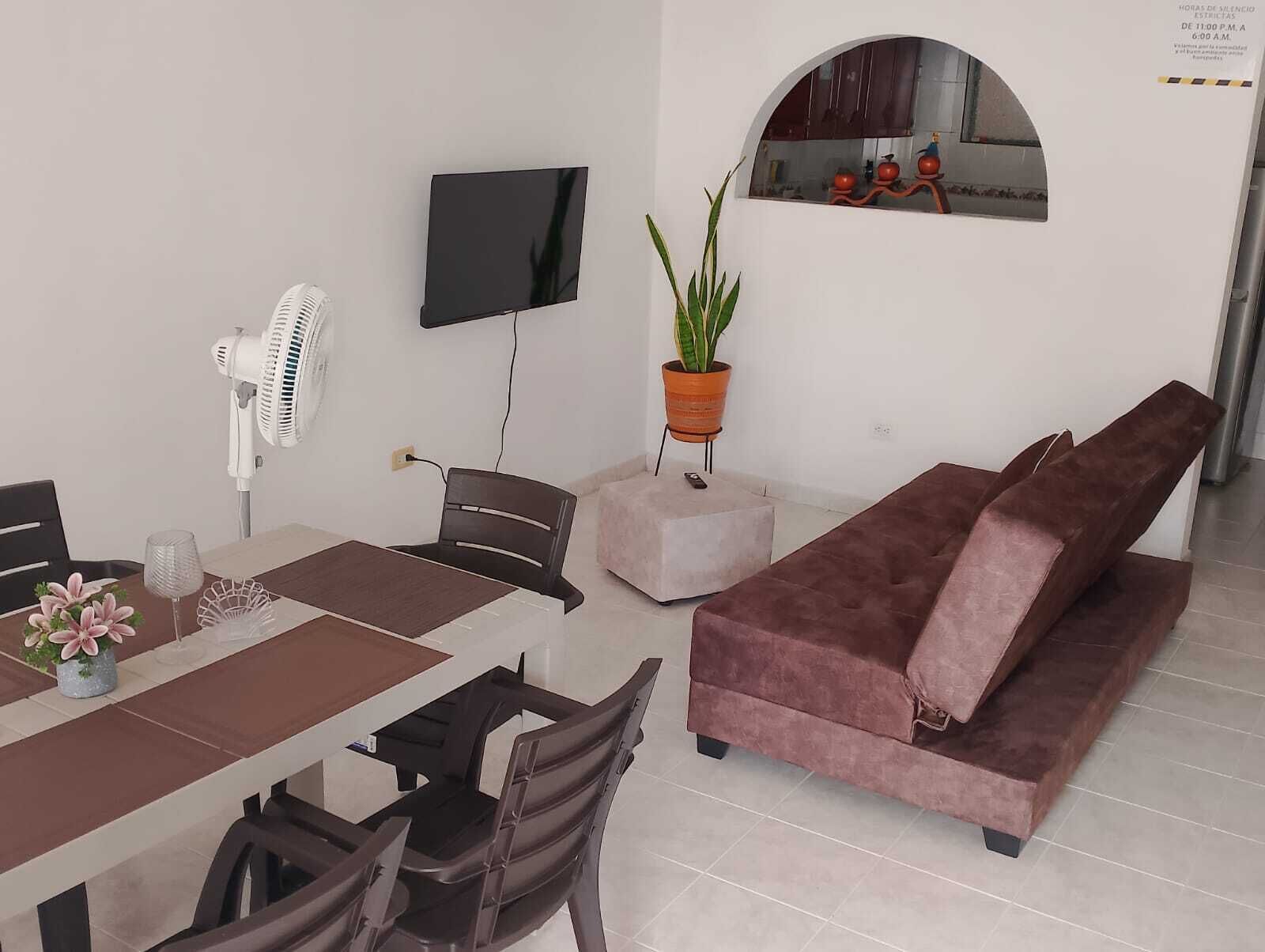 A modern living room with a dining area, a flat-screen TV mounted on the wall, a potted plant, a sofa, and a fan.